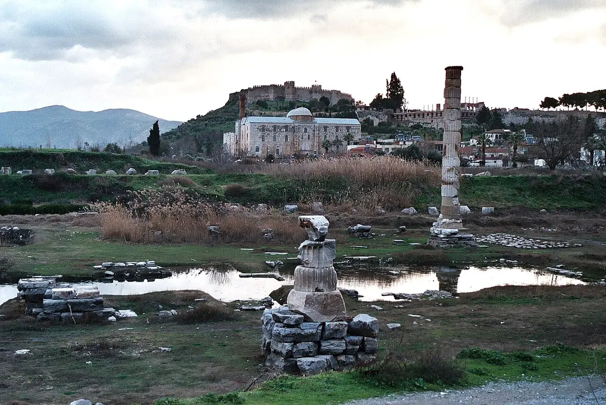 A single reconstructed column standing among scattered stone blocks and marshy water at the site of the Temple of Artemis, with a hilltop castle and mosque in the distance