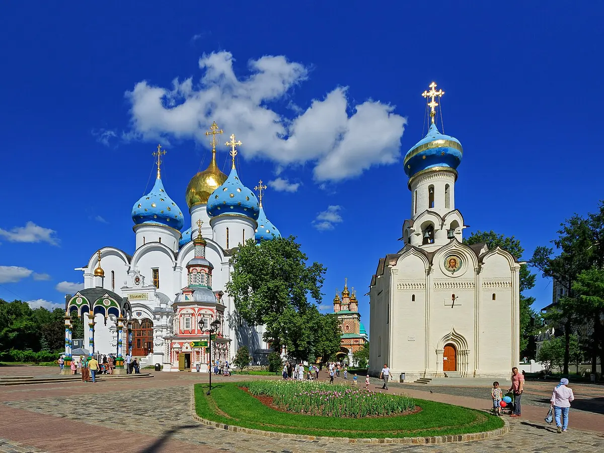A white church with a single blue dome and a bell tower on the right, with the blue and gold domes of a large cathedral behind, under a blue summer sky