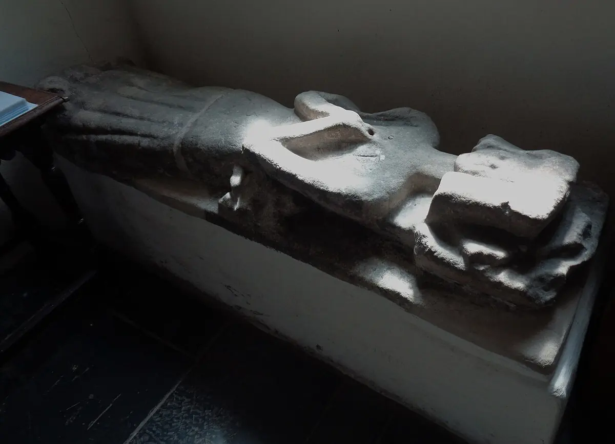 A worn medieval stone effigy of a robed woman lying on a low tomb chest in a dim corner of a church