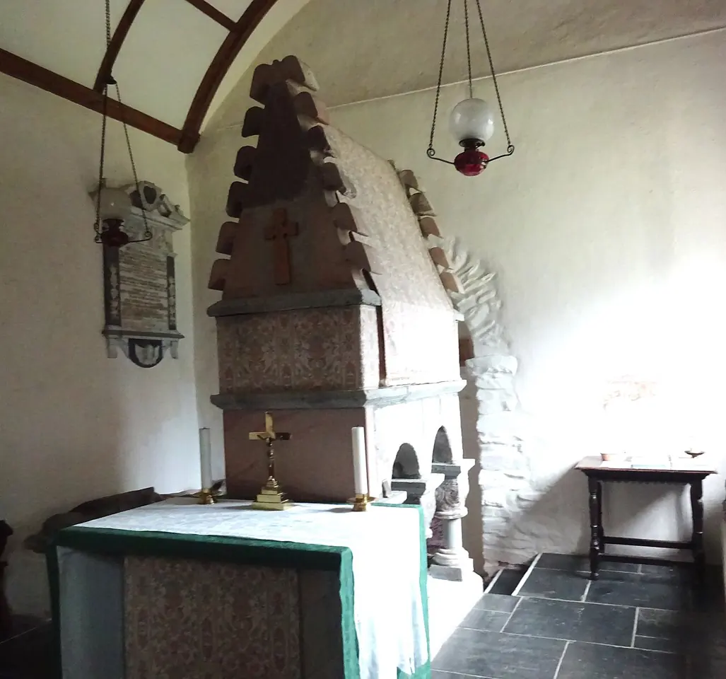 A tall reconstructed stone shrine with a gabled, crested top and a carved cross, standing behind a small altar in a whitewashed apse