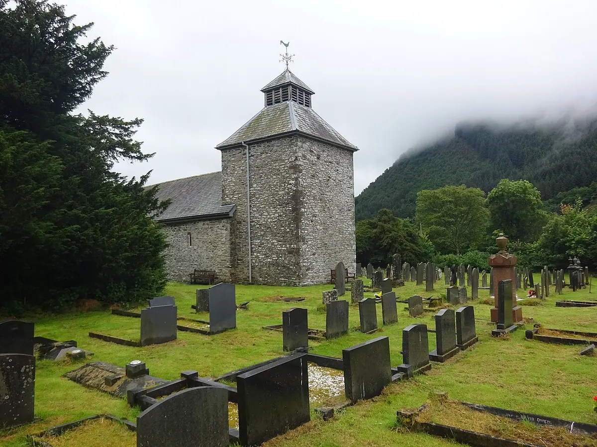 A small gray stone church with a square tower topped by a little belfry, in a churchyard of slate headstones under huge dark yew trees and misty green hills