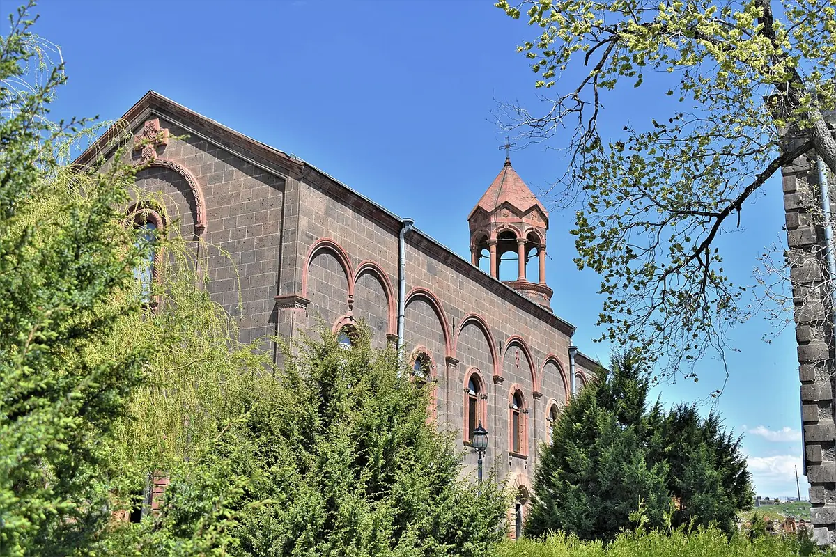 A long dark-stone church with tall arched windows and a small bell tower with a conical roof, seen through green trees under a blue sky