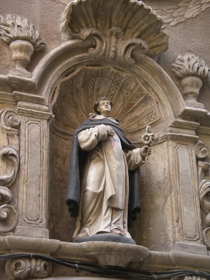 Painted stone statue of a young friar in a white habit and dark cape holding a small monstrance, standing in a carved shell niche