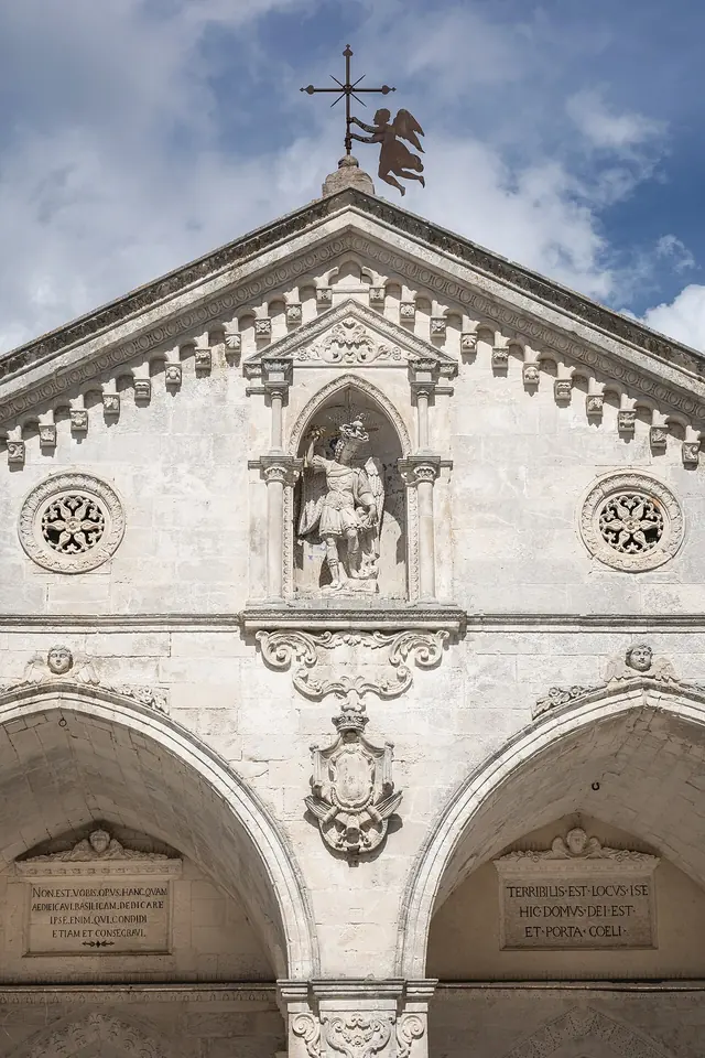 Fachada gótica de piedra blanca del santuario de Monte Sant'Angelo, con dos arcos apuntados, rosetones y una estatua de San Miguel en un nicho bajo el frontón