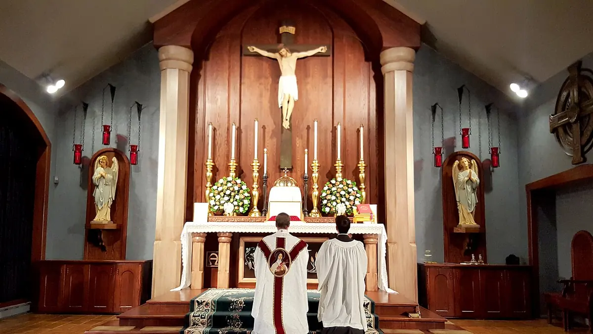 A priest in a white chasuble and a server in a surplice kneel at the foot of an altar with six candles and a large crucifix, between statues in side niches