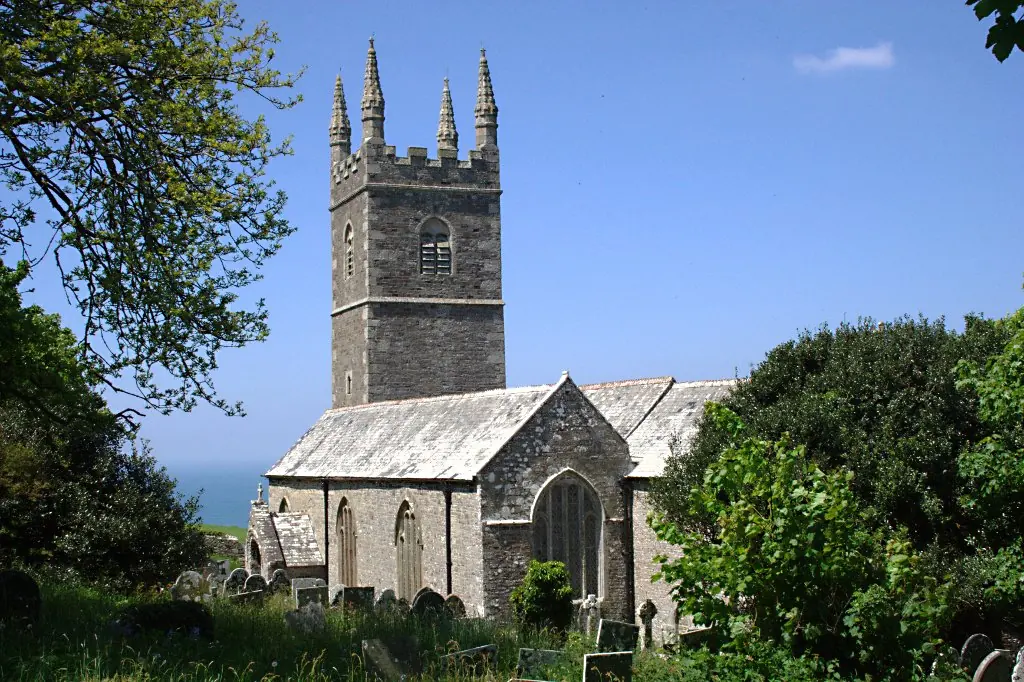 A gray stone church with a tall pinnacled tower among trees and gravestones, with a glimpse of the sea beyond under a blue sky