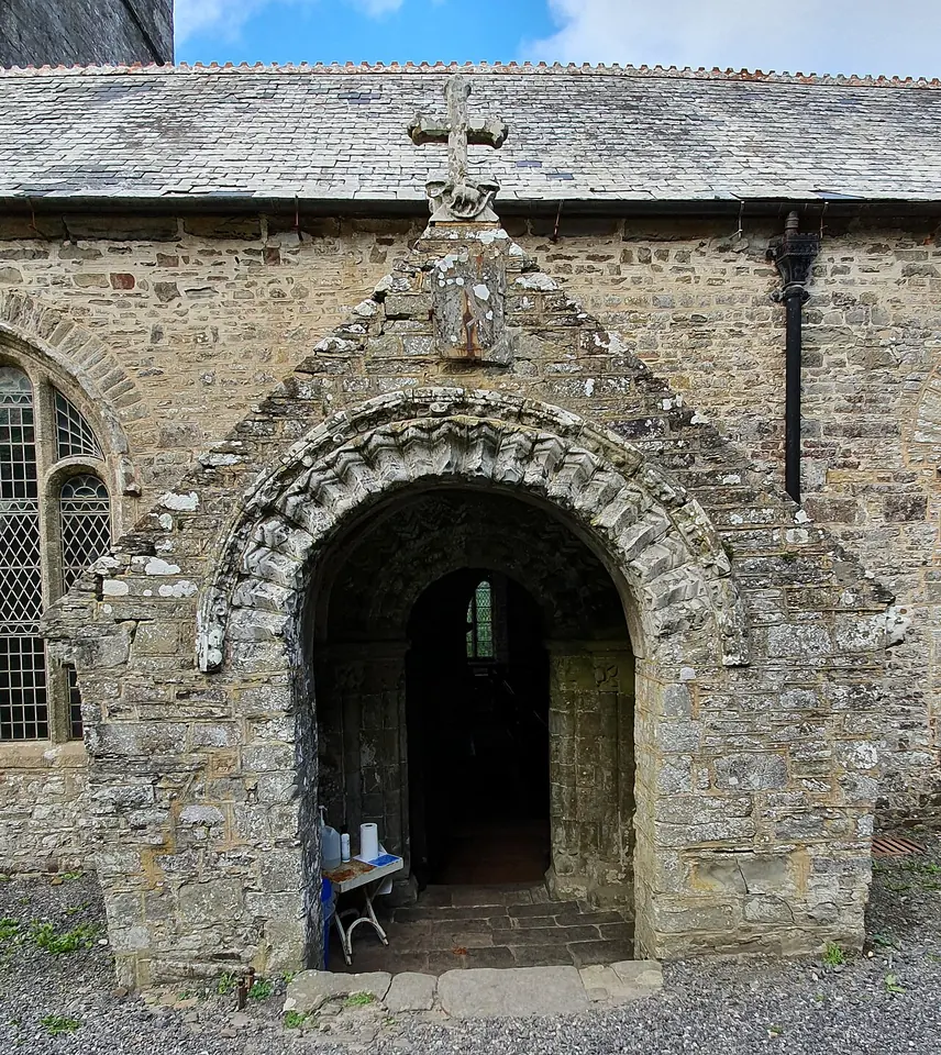 A stone church porch with a round arch carved in zigzag patterns, a small cross on its gable and a carved lamb above the doorway