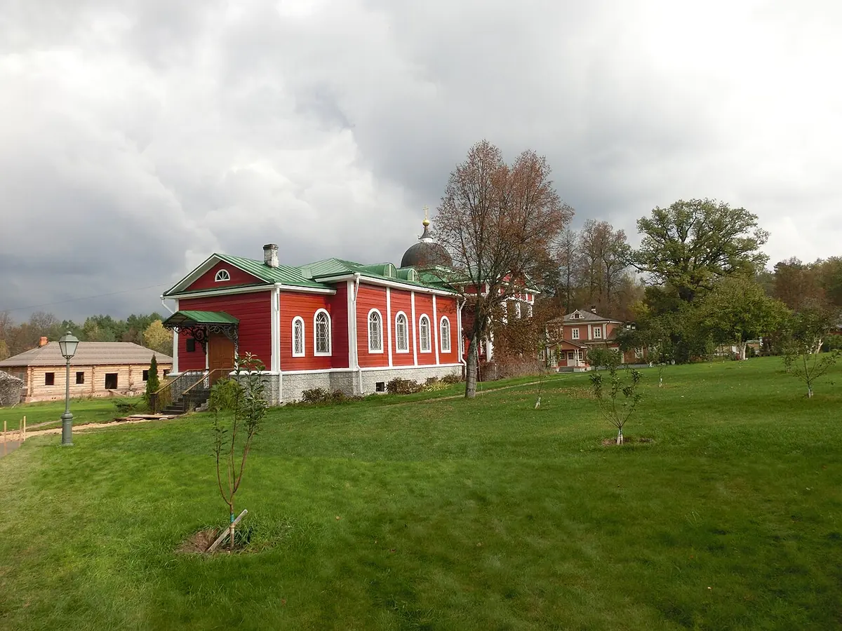 Small red wooden church with white window frames and a green roof and dome, standing on a wide green lawn under a cloudy sky