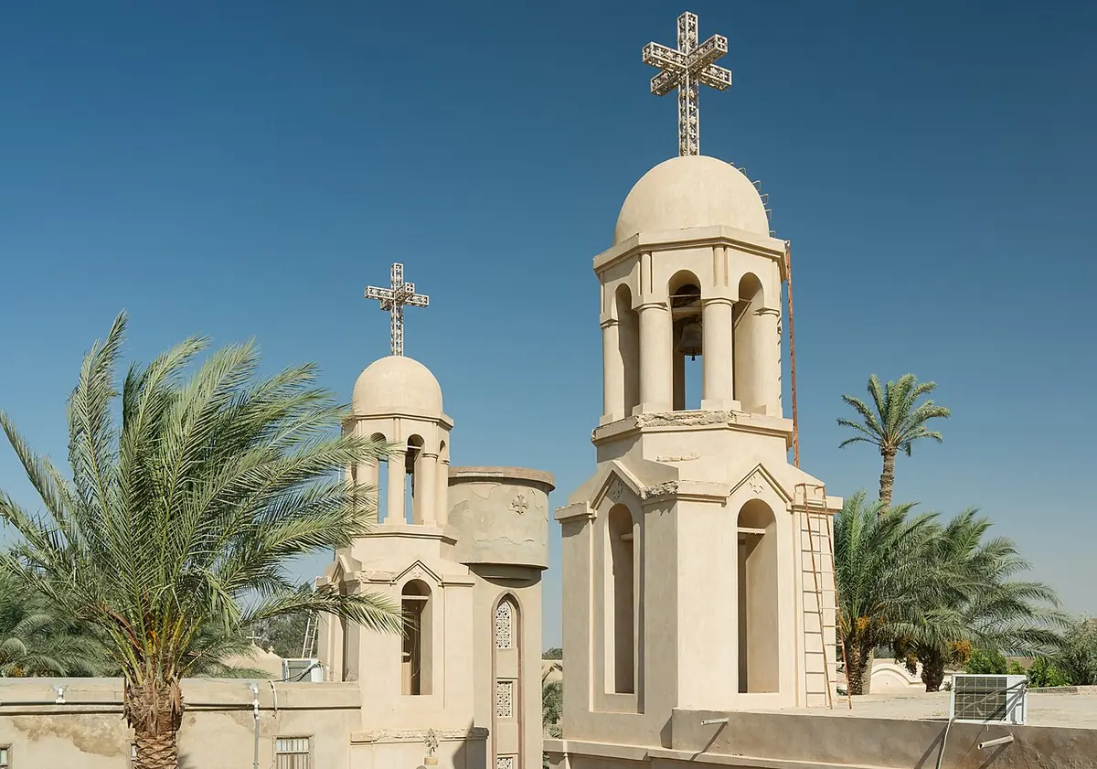 Two white domed bell towers topped with crosses rising above palm trees against a blue sky at a Coptic monastery in the Egyptian desert