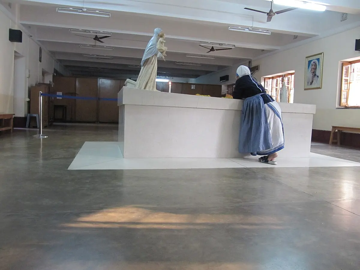 A sister in a white sari bends over a plain white marble tomb in a bright, bare room, arranging flowers, with a statue of the Virgin at the far end