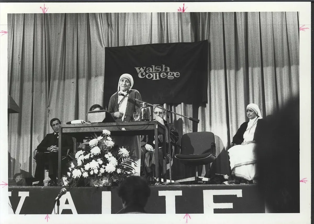 Black-and-white photograph of Mother Teresa speaking at a lectern on a stage under a Walsh College banner, with another sister seated beside her