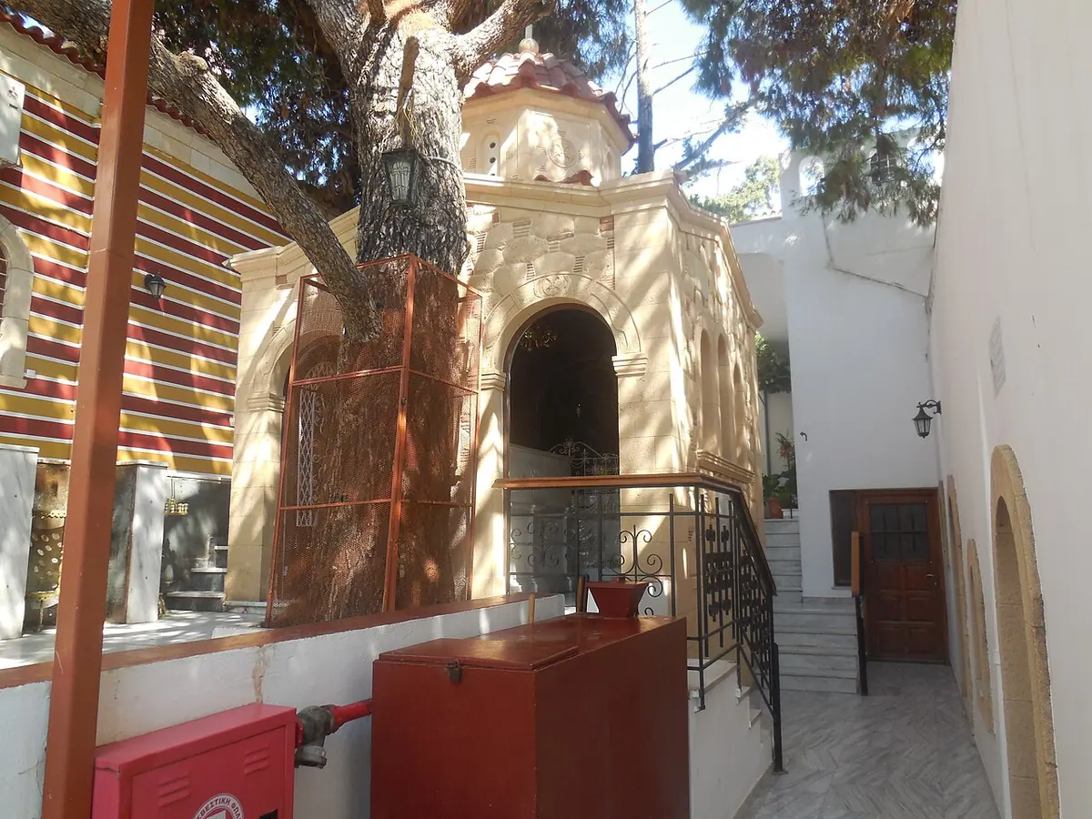 A small domed white chapel under a large pine tree in a monastery courtyard, beside a striped church wall