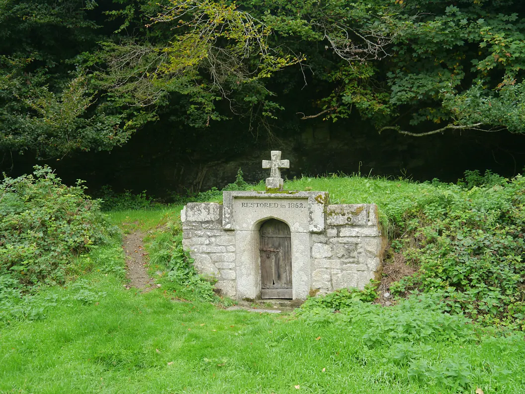 Small stone well-house with a wooden door and a cross on top, set into a grassy bank below trees