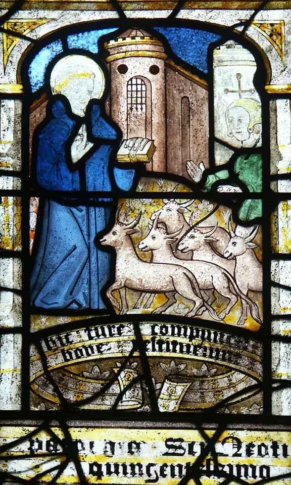 Medieval stained-glass panel of a haloed monk in a blue habit holding a book, with a group of pale stags in front of him and a towered building behind, above a scroll of black-letter text
