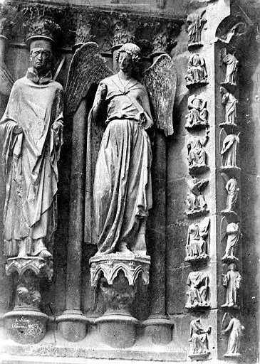 Old black-and-white photograph of two Gothic statues on a cathedral portal, a bishop on the left and a winged, smiling angel beside him