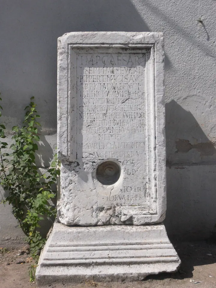 Weathered white stone Roman monument with a carved Latin inscription standing against a wall