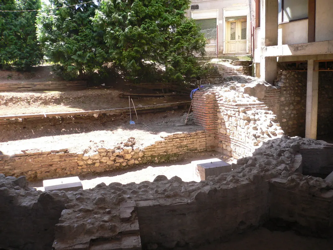 Excavated foundations of thick Roman stone and brick walls in a dug-out site between modern buildings and trees