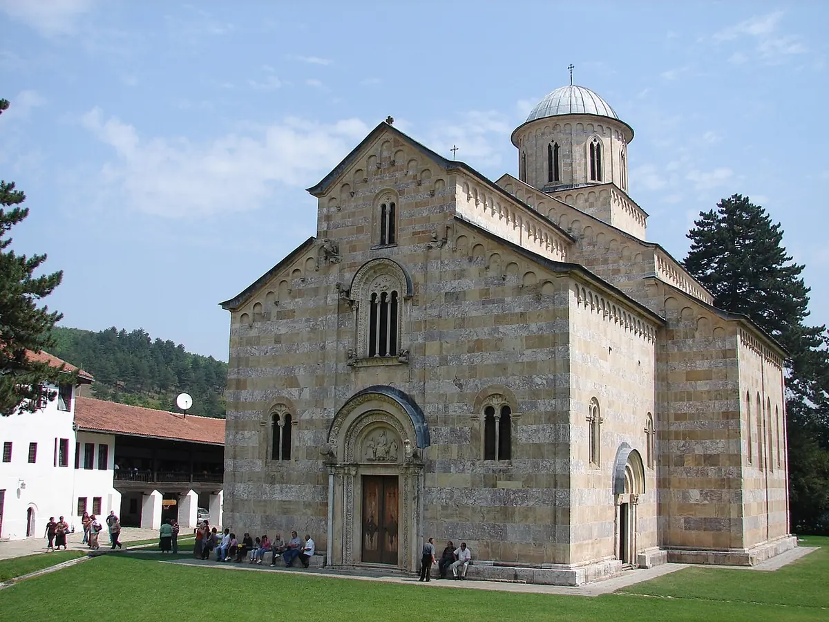 A large medieval stone church with banded marble walls, a central dome and a carved portal, on a lawn with pilgrims, in front of white monastery buildings and green hills