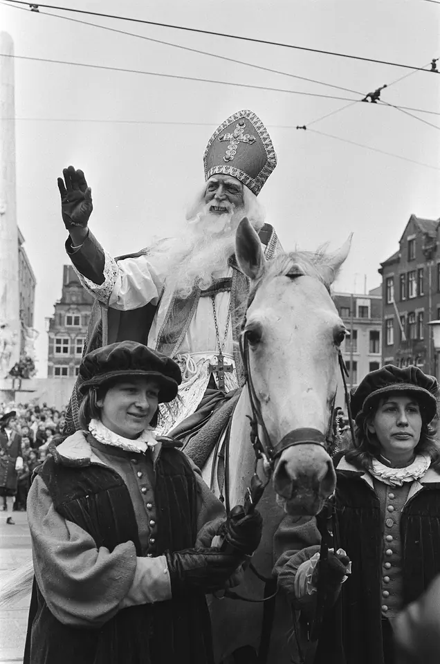 Black-and-white photograph of Sinterklaas in a tall miter and white beard waving from a white horse, led by two helpers in velvet caps and ruffs