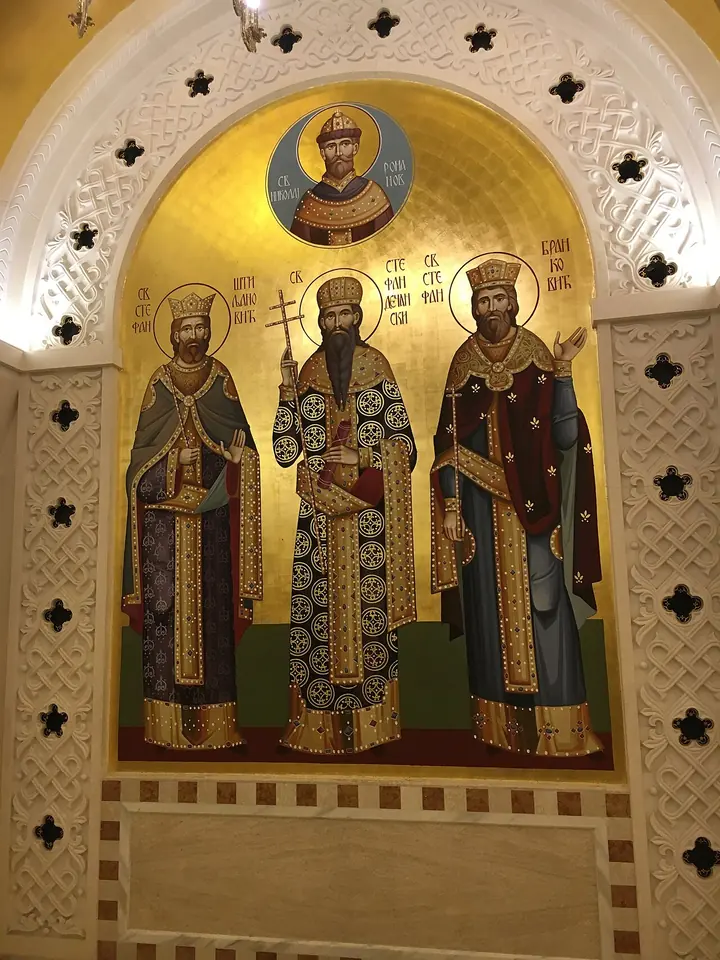 A gold mosaic in an arched niche showing three crowned Serbian saints in robes, with a round medallion of Tsar Nicholas II above them