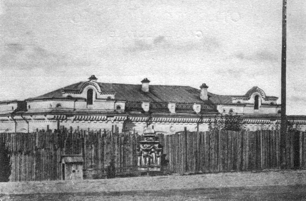 Old photograph of a two-story house behind a tall wooden palisade fence on a sloping street