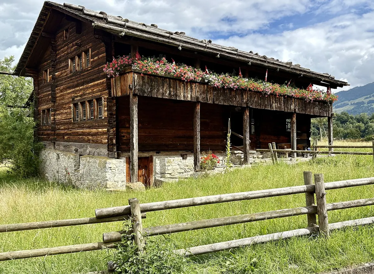 A large old Swiss farmhouse of dark weathered timber on a stone base, with a long balcony hung with red geraniums, behind a rail fence in a green meadow