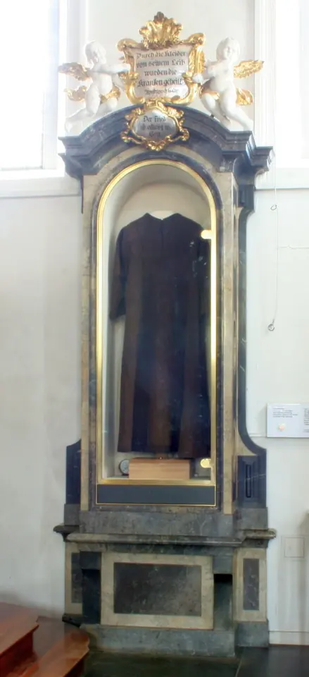 A dark brown woolen habit displayed in a tall glass case set in a gray and gold Baroque frame against a white church wall