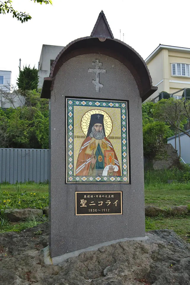 Gray stone memorial with a small roof and cross, holding an icon of St. Nicholas in bishop's vestments above a plaque reading Saint Nicholas, 1836 to 1912, in Japanese