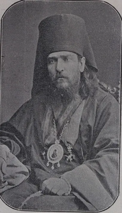 Black-and-white photograph of a bearded Orthodox bishop in a tall dark klobuk and mantle, wearing a panagia, seated with his hands folded