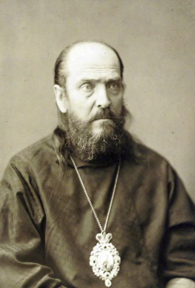 Sepia portrait photograph of an older balding bishop with a long gray beard, wearing a dark cassock and a large ornate panagia on a chain