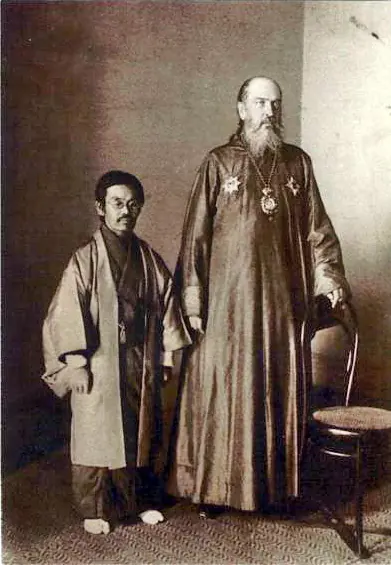 Old sepia photograph of a tall bearded Orthodox bishop in a long cassock standing beside a much shorter Japanese man in a kimono and haori