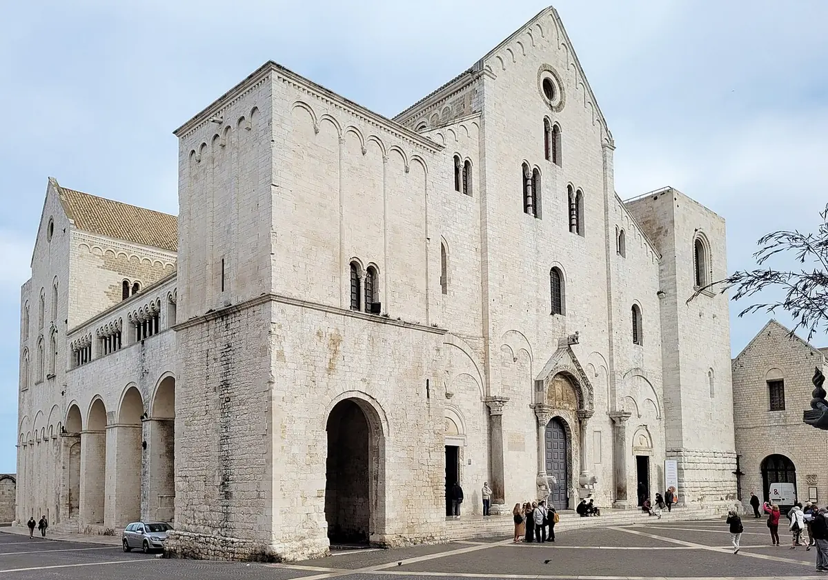 Fachada románica de piedra clara de la basílica de San Nicolás de Bari, con dos torres bajas y una arcada lateral, vista desde la plaza