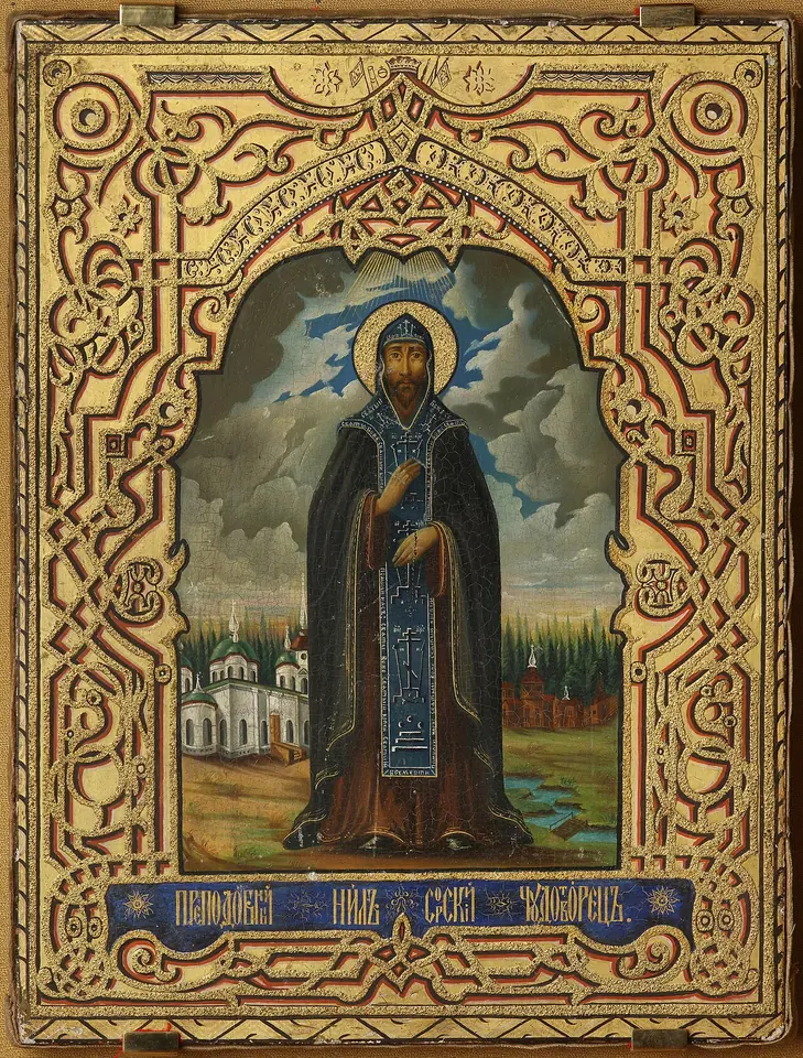 Painted icon of a monk in a black schema holding a prayer rope, white monastery churches behind him on one side of a river and wooden buildings on the other, in an ornate gilded border