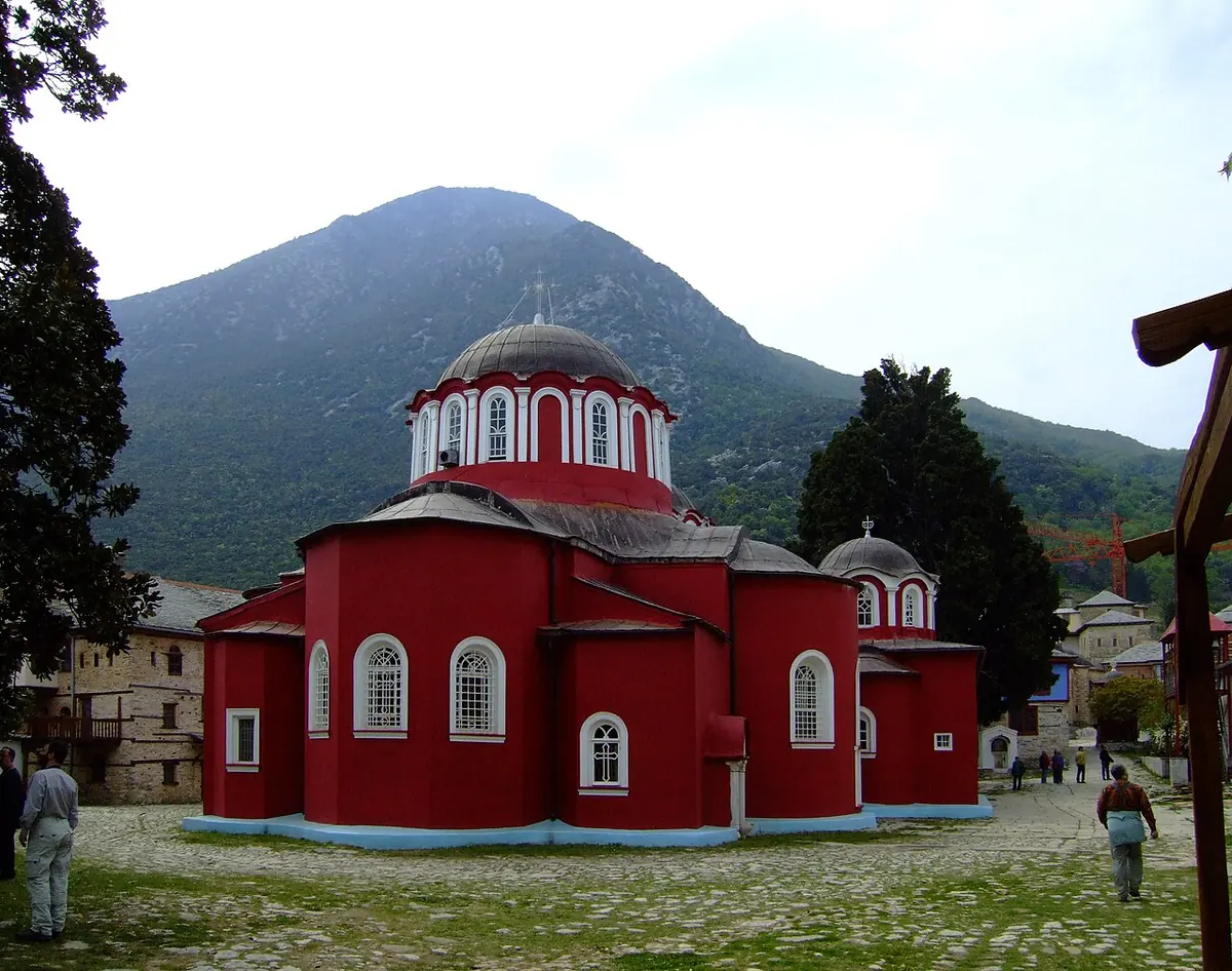Large red-painted Byzantine monastery church with several domes in a cobbled courtyard, a wooded mountain rising behind it