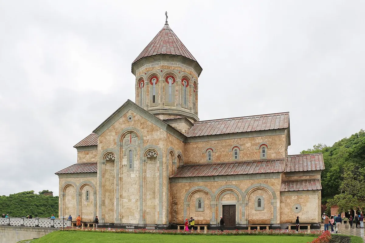 A Georgian stone church with a central drum and conical dome, arched windows and a long tiled roof, set on a lawn under a cloudy sky