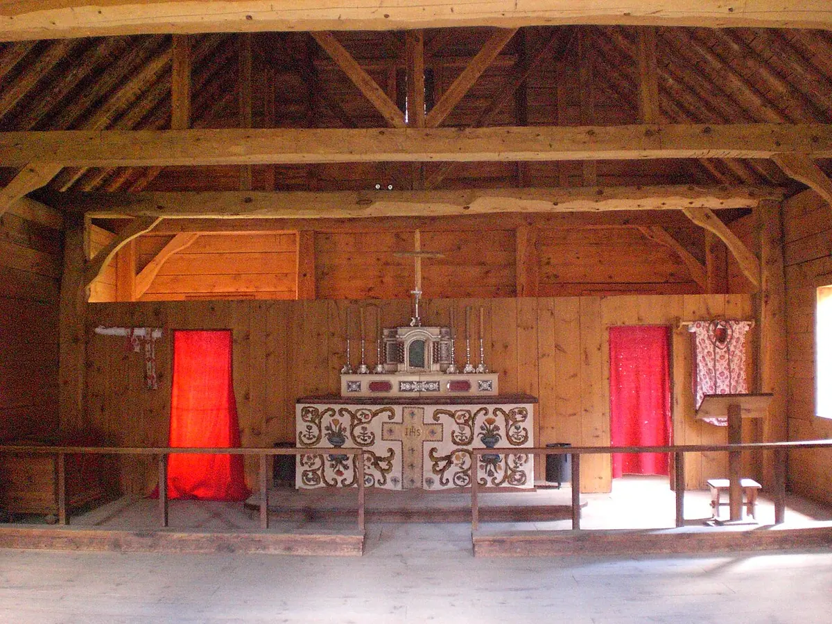 Interior of a timber chapel with a plain wooden altar, a cross, red hangings and log walls