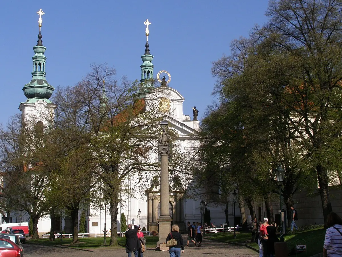 The white Baroque facade and twin green-domed towers of the Strahov abbey church in Prague, seen across a tree-lined courtyard with a column statue of St. Norbert