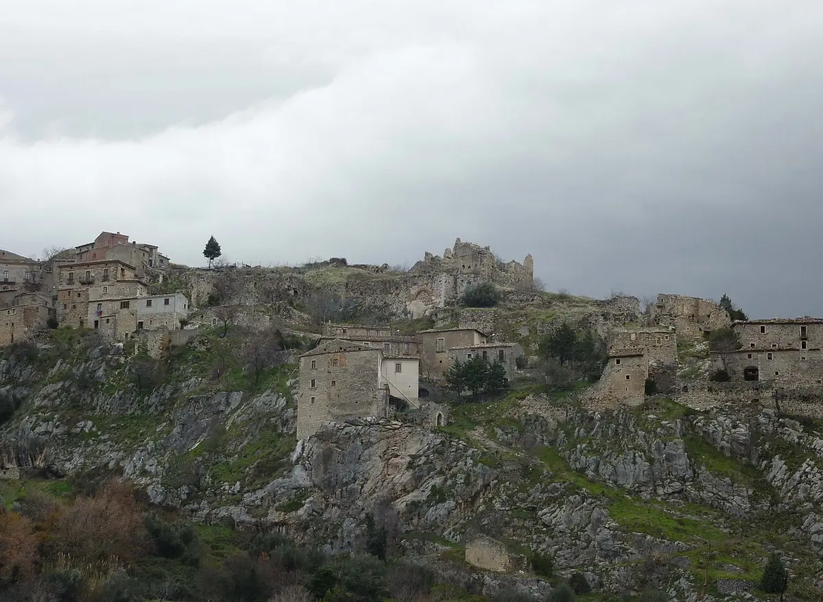 Stone houses and a ruined castle along a rocky ridge above green slopes under a gray sky
