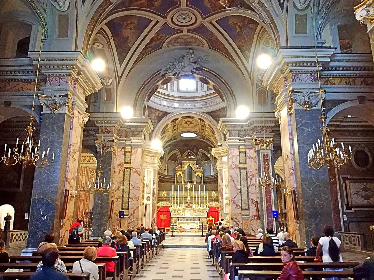 The nave of a Baroque church with colored marble columns, a painted dome and a gilded high altar, with people seated in the pews