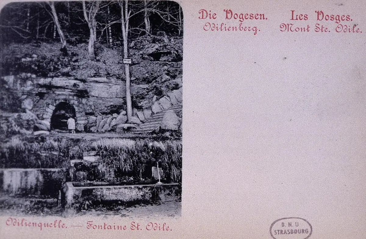 An old postcard photograph of a small stone spring house under trees on a wooded hillside
