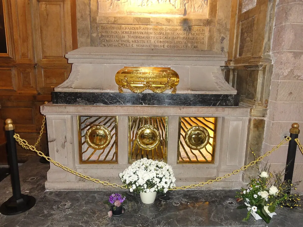 A stone sarcophagus with a gilded reliquary on top, flowers at its foot and a carved inscription on the wall behind