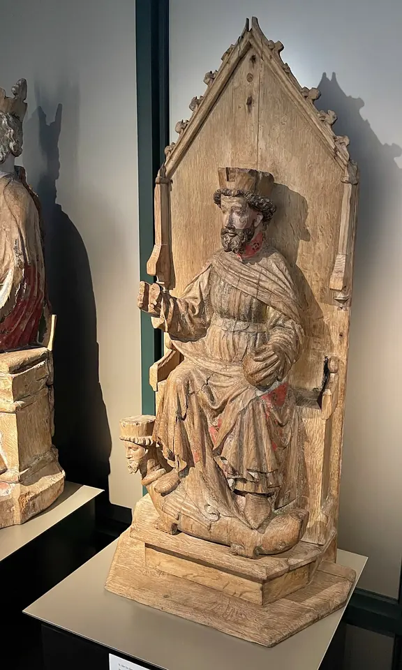 Unpainted wooden statue of a crowned, bearded king seated on a Gothic throne with his feet resting on a beast with a human head