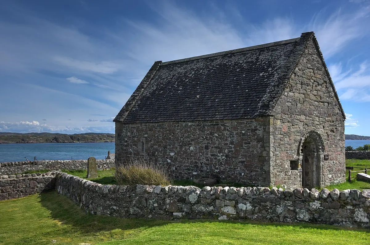 Small gabled medieval stone chapel with a round-arched doorway, surrounded by a dry-stone wall and grass, with the sea behind it