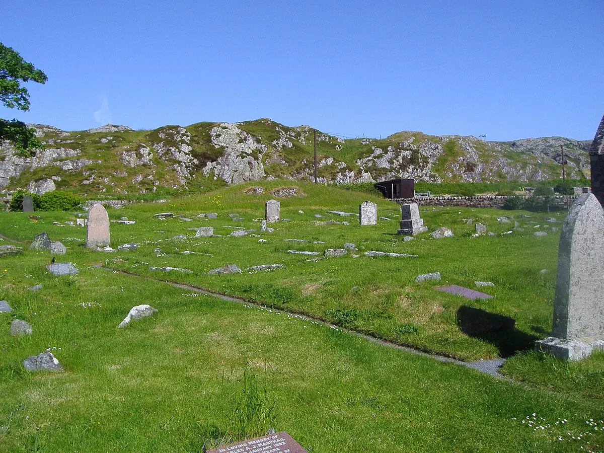 Green graveyard with scattered old headstones and grave slabs below a rocky hillside under a clear blue sky