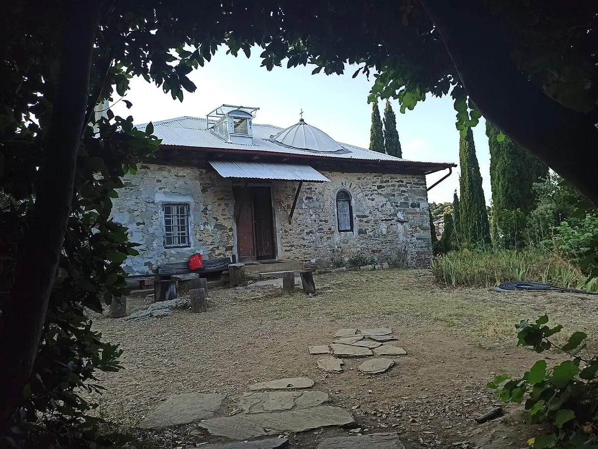 A low stone hermitage house with a gray roof and an awning, seen through overhanging branches across a courtyard of flagstones and dirt