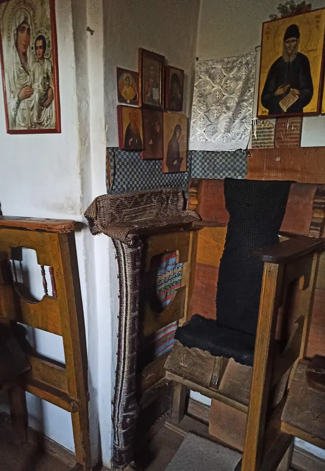 A worn wooden church stall with a dark cushion in a small whitewashed chapel, with icons of the Virgin and of St. Paisios on the wall above it