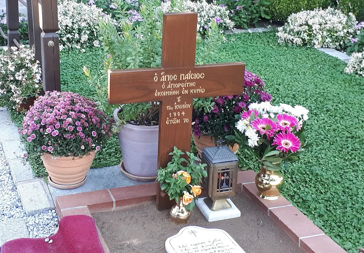 A simple grave with a wooden cross, an oil lamp, potted flowers and a small white marble plaque, set in a lawn