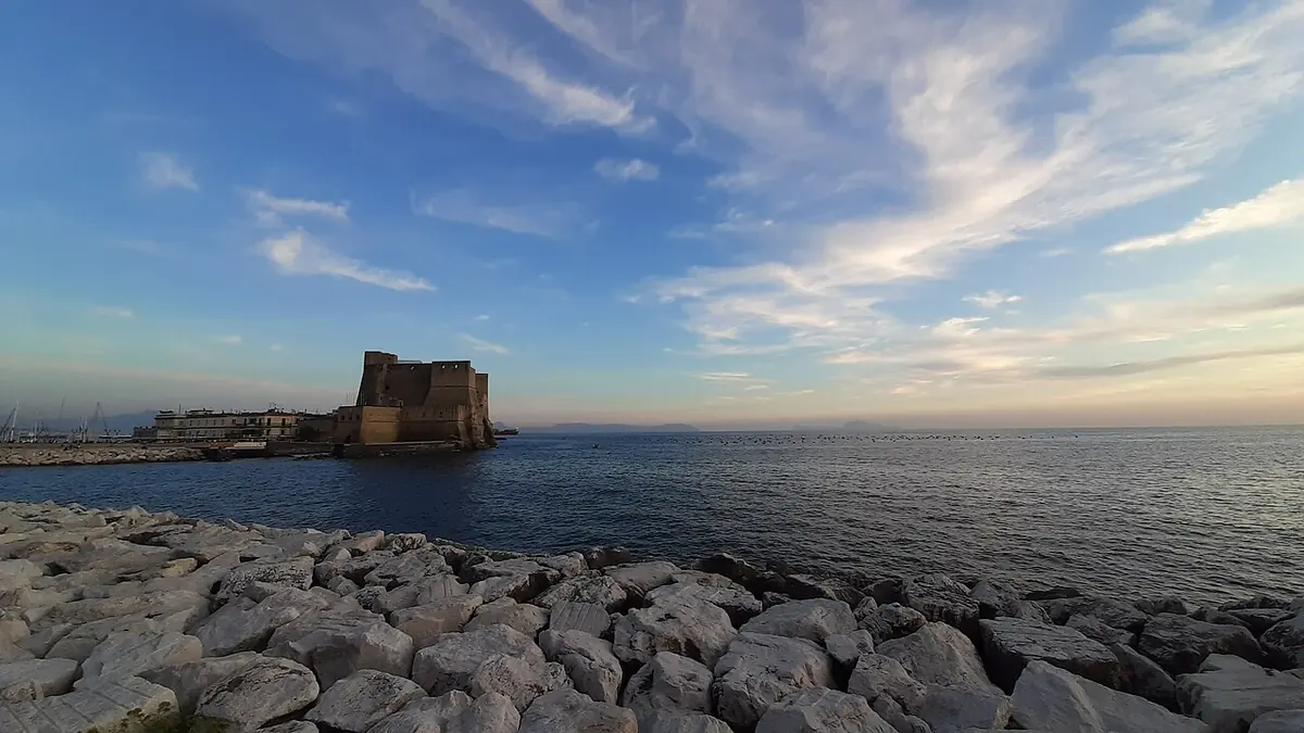 A low stone castle on a small islet at the end of a breakwater in a calm bay at sunset, with streaks of cloud across a blue sky