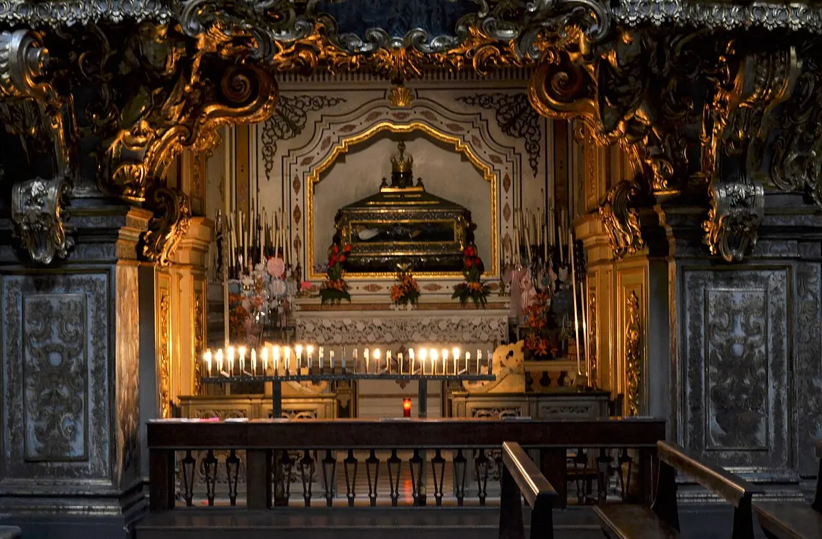 A richly gilded Baroque chapel with a glass urn high above the altar, candles burning before it and a wooden railing in front