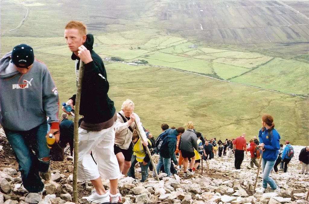 A long line of pilgrims with walking sticks climbing the loose stone path of Croagh Patrick, with green fields and the plain far below
